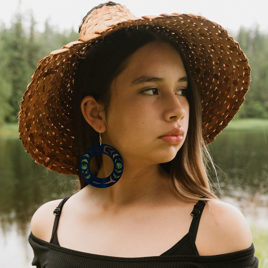 A woman stands by the water wearing a cedar hat and cobalt mirror acrylic Nala Hoop earrings from Copper Canoe Woman.