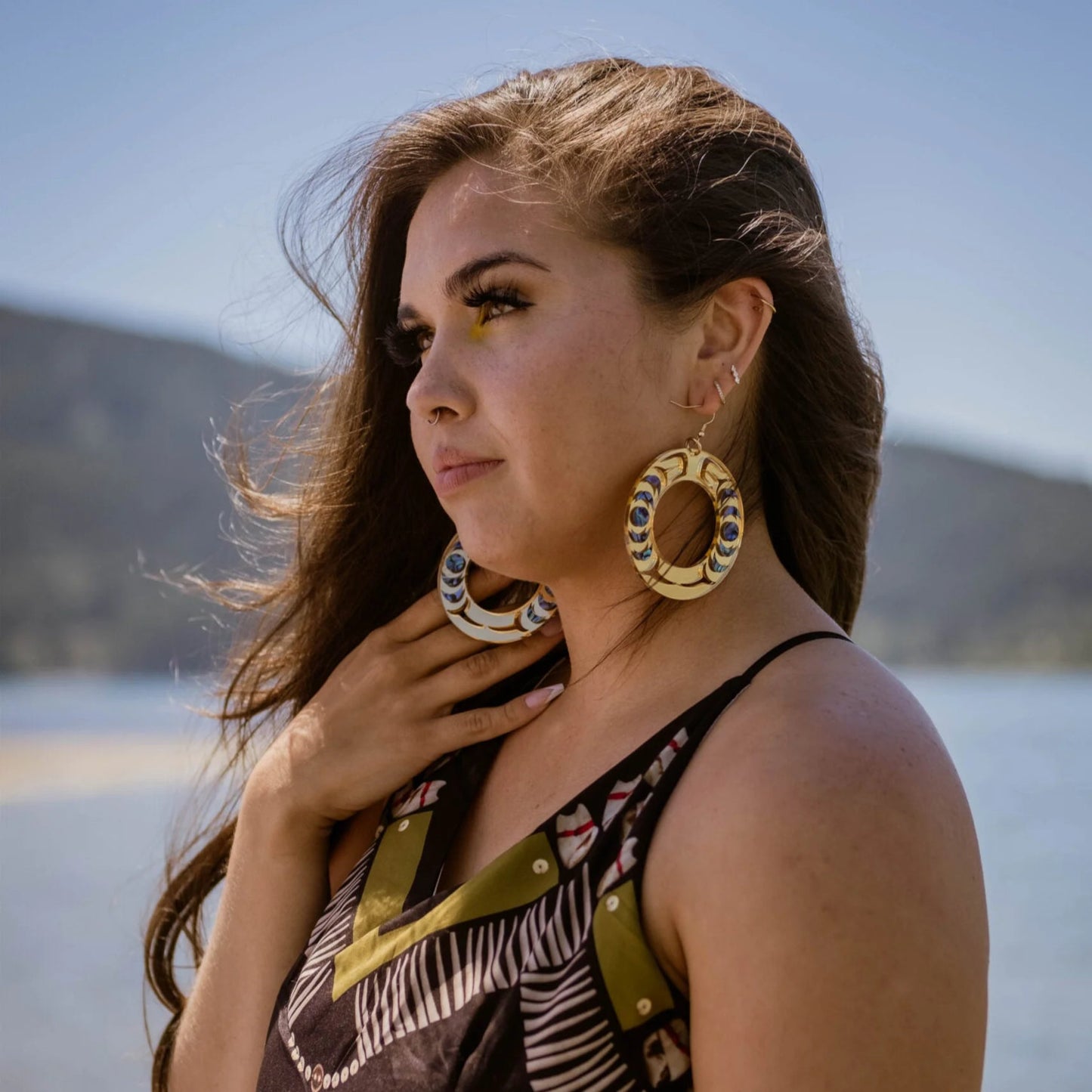 A model stares at the sea wearing the "Nala Hoop" earrings in gold mirror acrylic from Copper Canoe Woman. 