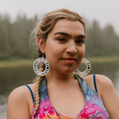 A model stares at the camera near the water wearing white acrylic Nala Hoop earrings with abalone in lay from Copper Canoe Woman.