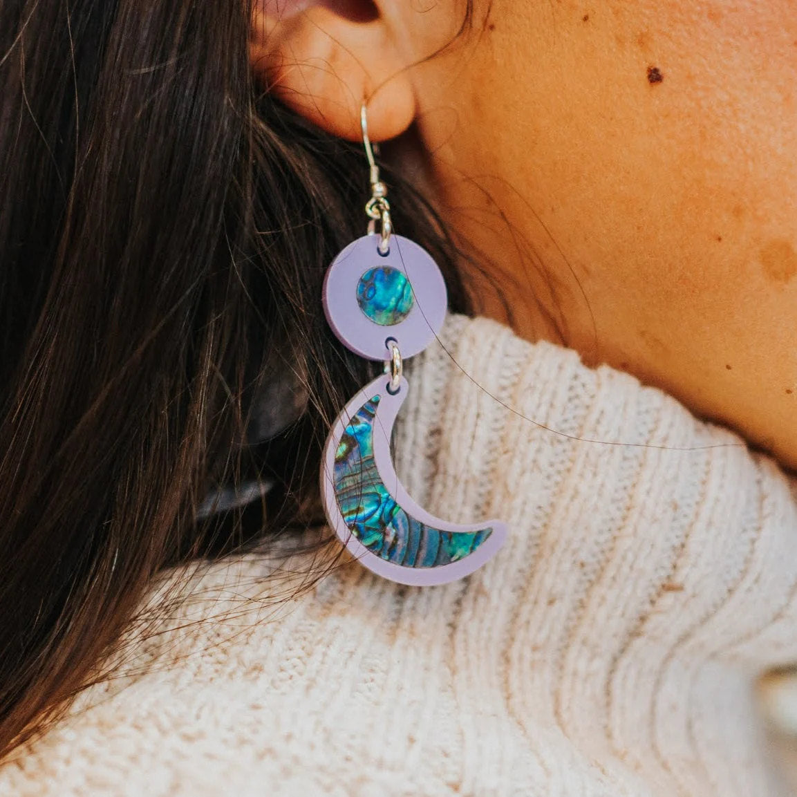 A closeup of a model wearing the hook "Nuci" crescent moon earring in lavender acrylic from Copper Canoe Woman.
