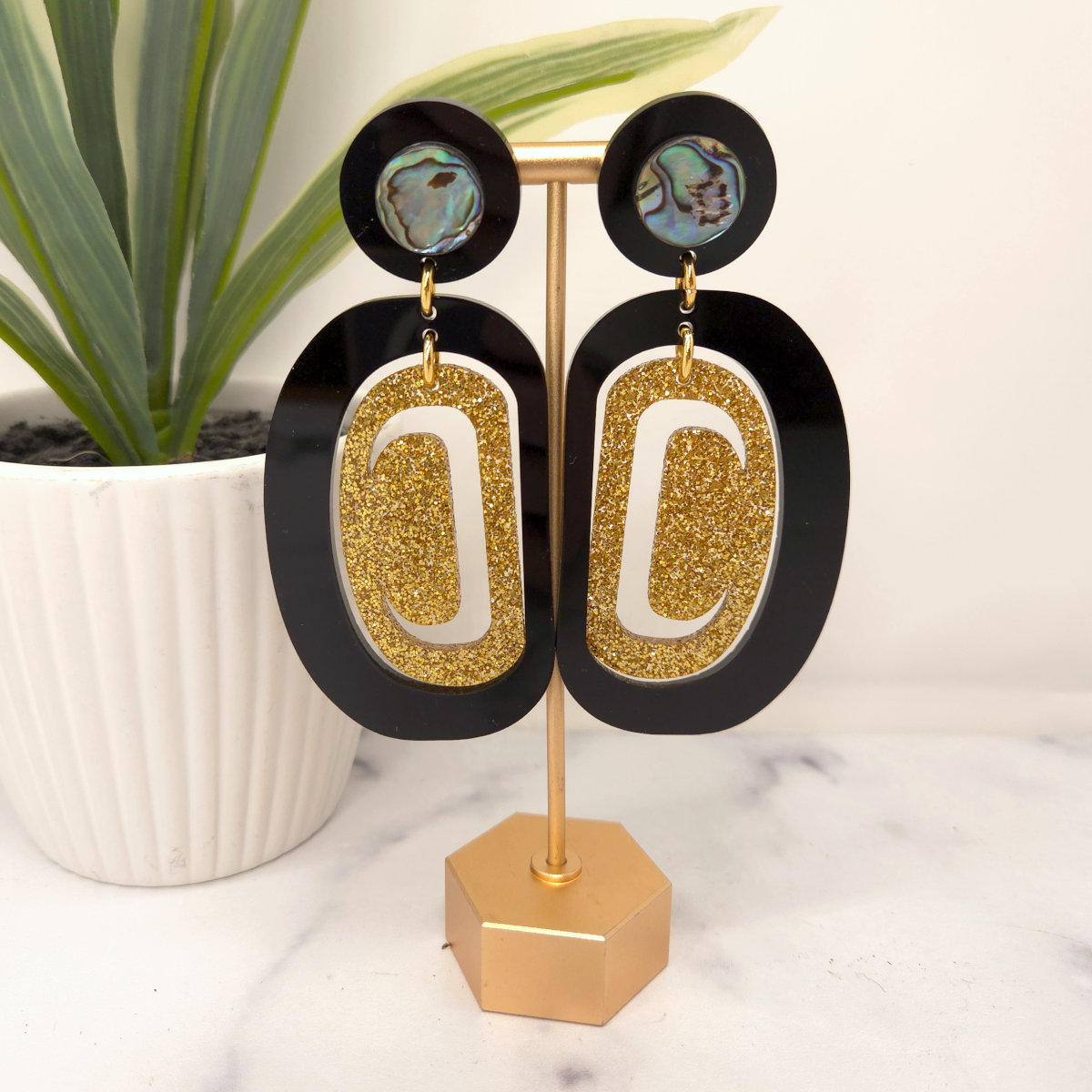 Gold and black earrings from Copper Canoe Woman on a stand with a plant in the background.