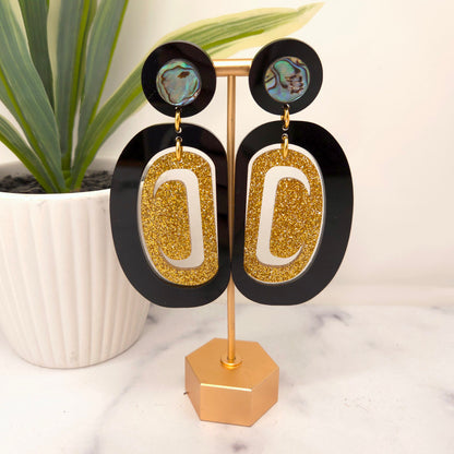 Gold and black earrings from Copper Canoe Woman on a stand with a plant in the background.