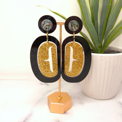 Gold glitter earrings from Copper Canoe Woman with black accents on a stand, plant in the background