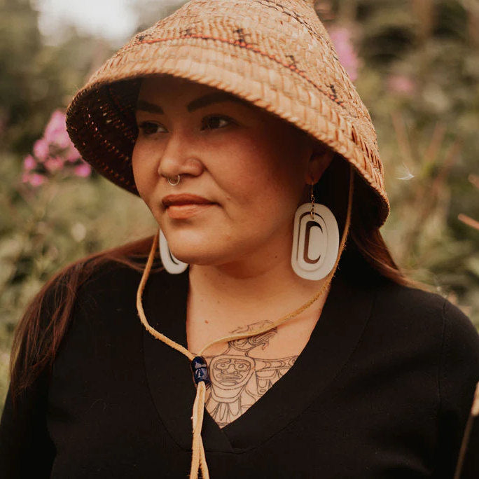 Person wearing a cedar hat and large ivory acrylic ovoid earrings with a blurred natural background