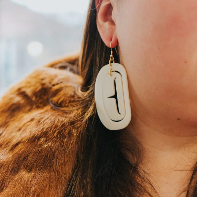 A close-up image of a person wearing a large ovoid ivory acrylic earring with a cutout design.