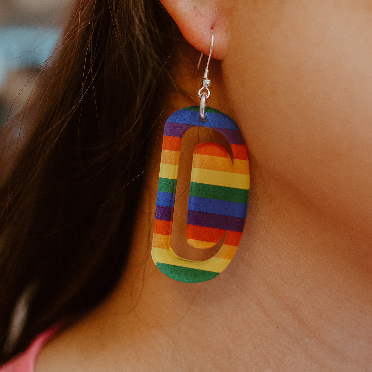 A closeup of a model wearing the small ovoid earring from Copper Canoe Woman in rainbow color acrylic.