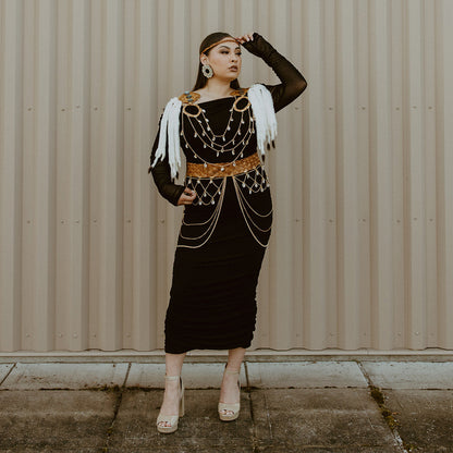 Woman in a black dress with gold accents standing against a beige wall.