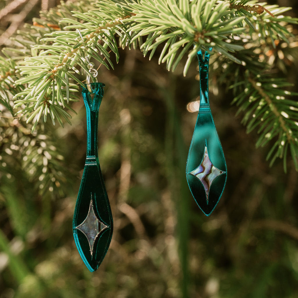 A pair of Copper Canoe Woman paddle-shaped earrings in teal mirror acrylic with abalone inlay hang from a pine branch.