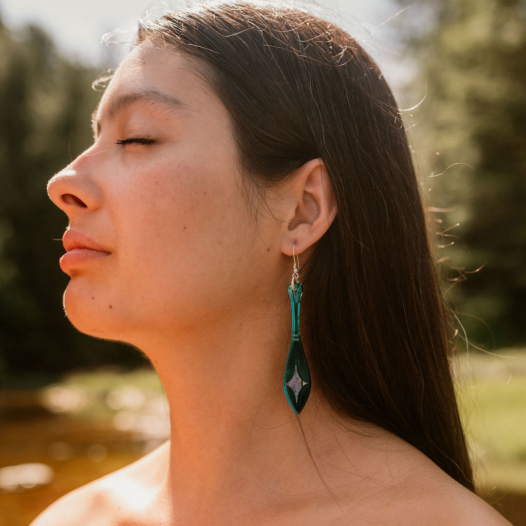 A profile of a woman wearing paddle shaped earrings in teal mirror acrylic with abalone inlay from Copper Canoe Woman.