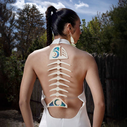 Woman wearing a white dress with cut-out copper canoe woman salmon ghost design on the back, standing outdoors.