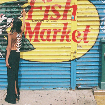 Woman with ivory copper canoe woman salmon ghost design on back of dress posing in front of a 'Fish Market' sign on a blue shutter.