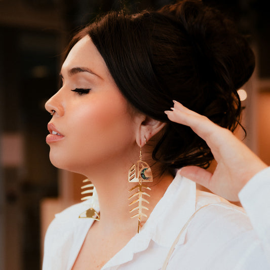 A person wearing a white shirt showcasing 14k gold-plated earrings with a salmon bone design from Copper Canoe Woman.