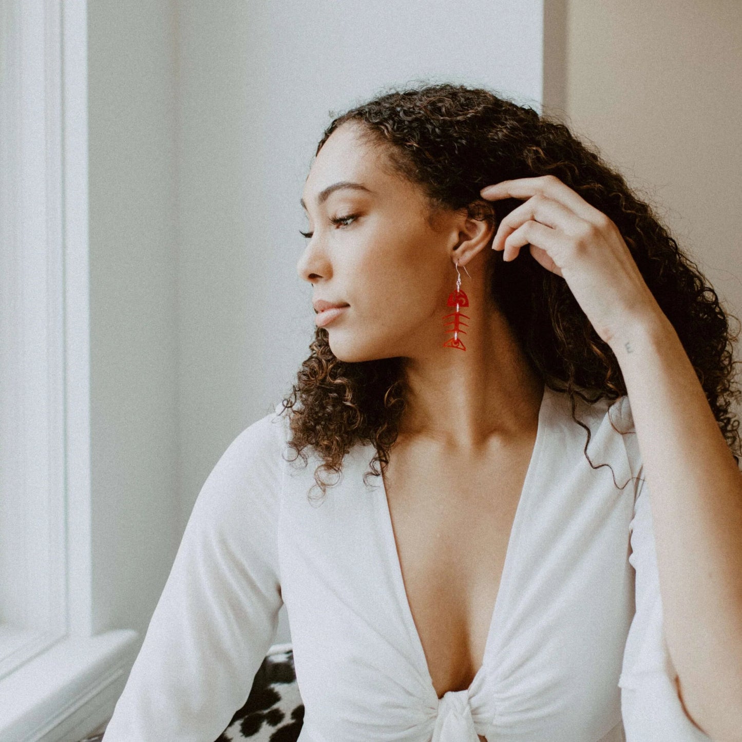 A woman looks out a window as she holds back her hair to show off the red acrylic small salmon ghost earring from Copper Canoe Woman.