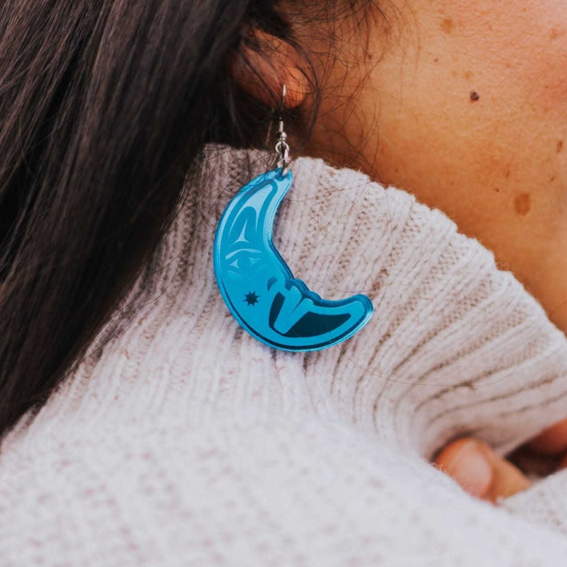 close up of woman wearing sapphire blue acrylic crescent moon engraved with formline art from copper canoe woman