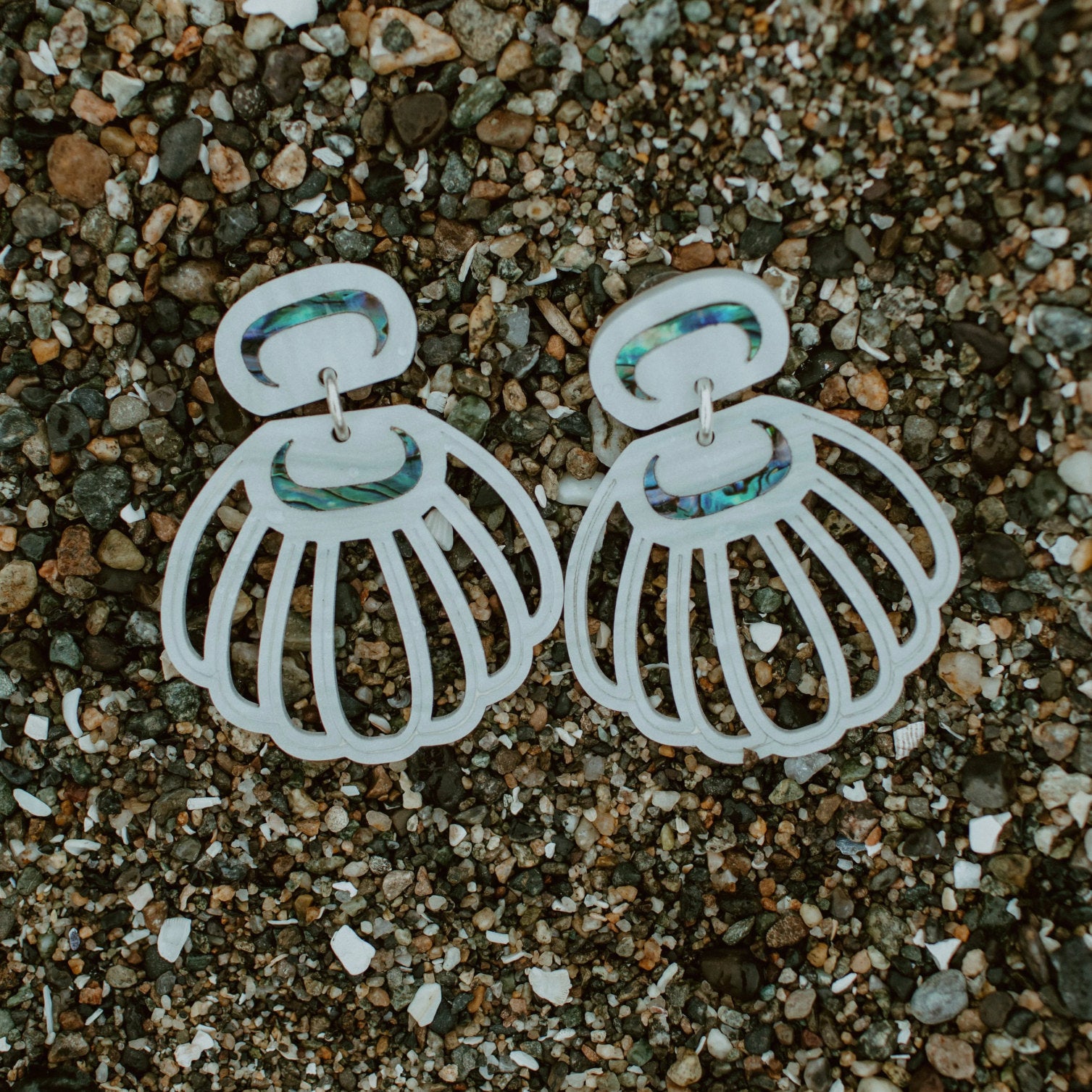 A pair of pearl acrylic "Shell Woman" earrings from Copper Canoe Woman rest on pebbles.
