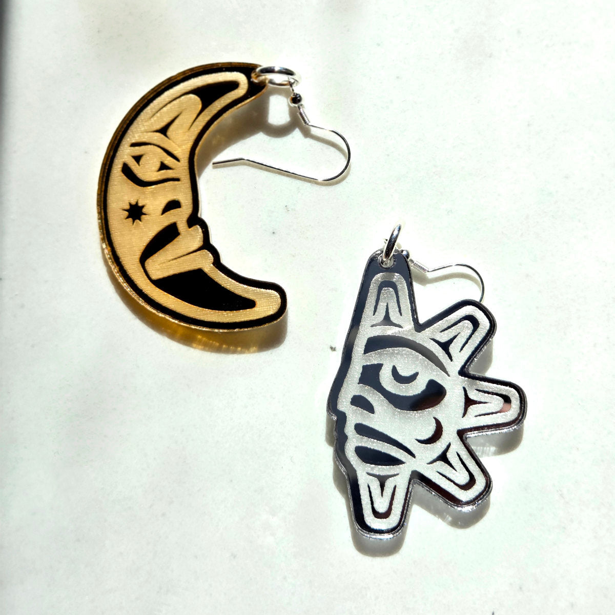 Two earrings, one sun-shaped and one moon-shaped with Coast Salish designs, on a white background from Copper Canoe Woman.