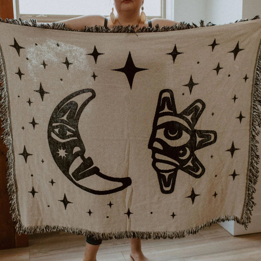 A figure holds open a "Hupal" sun and moon design throw blanket from Copper Canoe Woman.