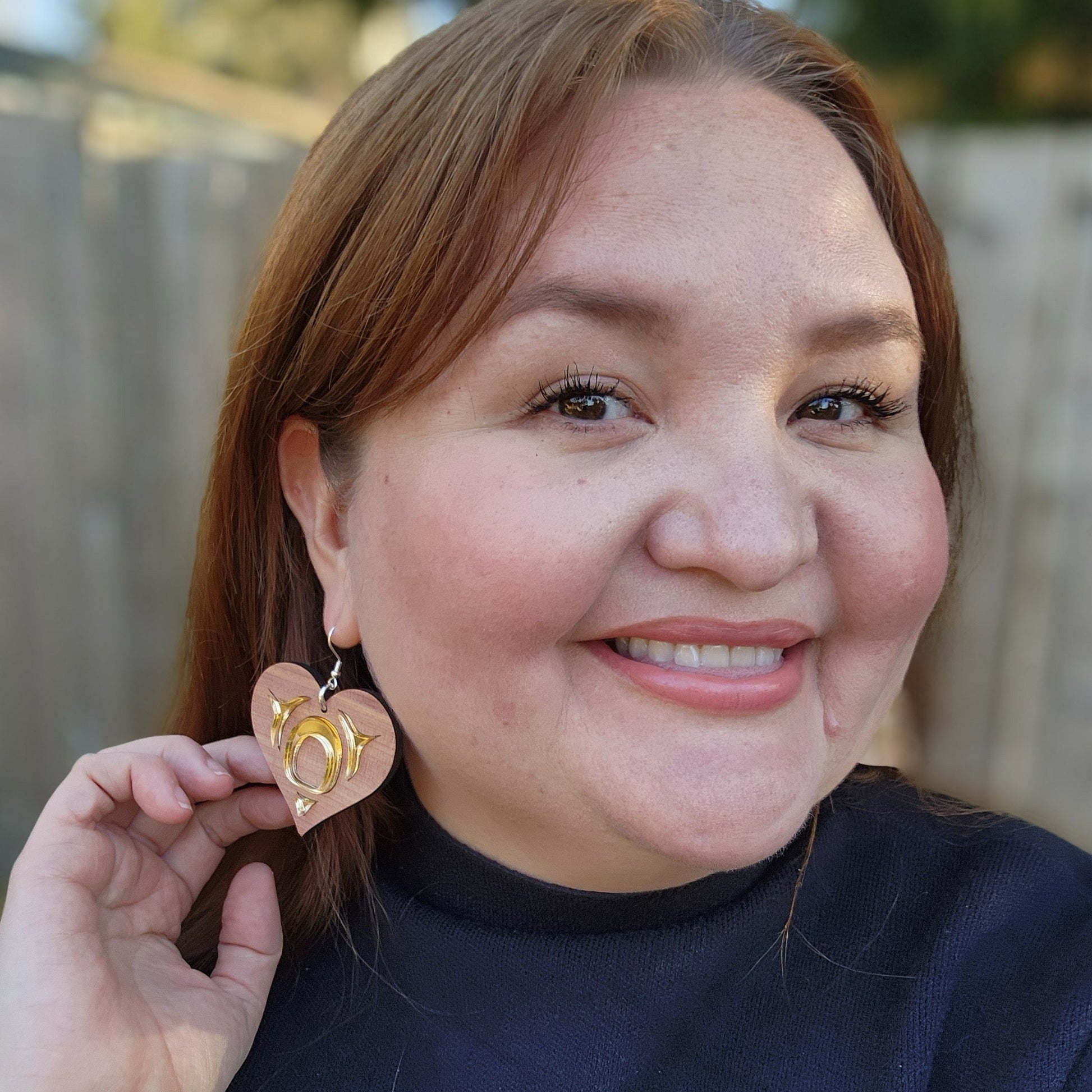 woman looking at the camera wearing copper canoe woman all my heart earrings in cedar with gold acrylic mirror formline art. 